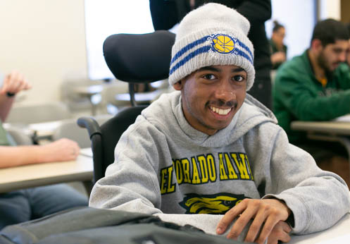 A smiling black male student sitting in his wheel chair at his desk.