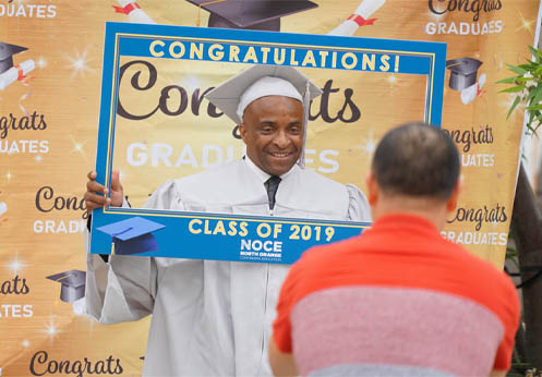 A student in their grey cap and gown smiling and holding up a sign