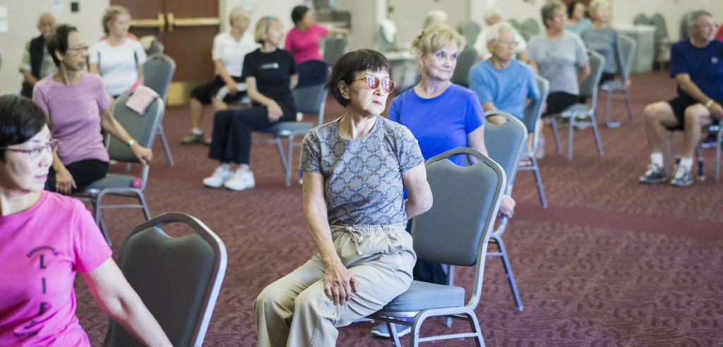 workout class with older students sitting on chairs performing stretches.