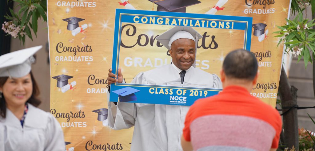 A photo of a student in their grey cap and gown smiling and holding up a “Congratulations Class of 2019” sign.