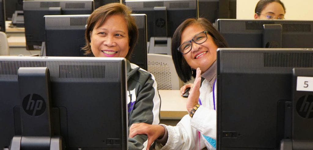 A photo to two students posing and smiling behind their computers monitors in classes.