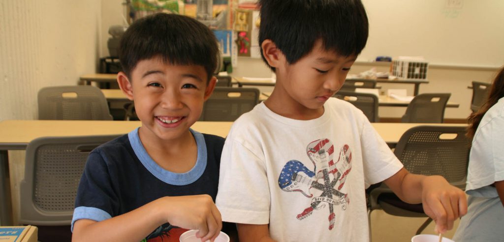 A photo of two students in class, both children. One is smiling for the camera, while the other kid is working on a project.