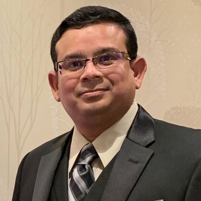 Dr. Riffat Ahmend professional headshot wearing glasses and dark suit with striped tie